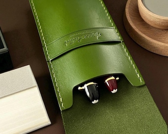 May include: A forest green leather pen case featuring a horizontal strap embossed with the text Aegean Leather. The case has visible light-coloured stitching along the edges and holds two pens with black and maroon caps visible at the top opening.