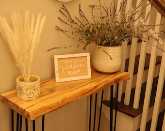 May include: A wooden console table with black hairpin legs, decorated with a white vase filled with lavender and a white sign that reads "Snuggle Season" and a white ceramic vase with dried pampas grass.