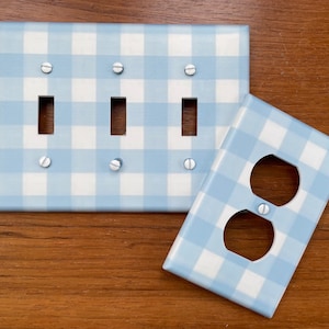 May include: Two light blue and white gingham patterned wall plates. One plate has three light switch openings and the other has a single outlet opening.