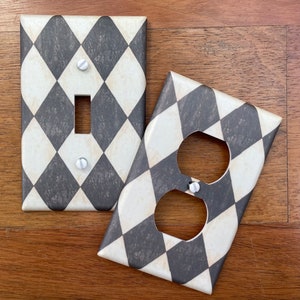 May include: Two decorative wall plates with a black and white harlequin pattern. One plate is a single light switch cover and the other is a double outlet cover.