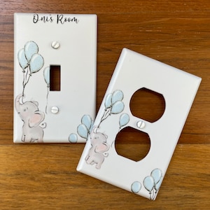 May include: Two white light switch and outlet covers with a light blue and gray elephant design. The switch cover has the text "Oni's Room" on it.