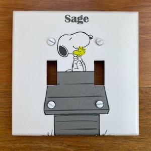 May include: White light switch cover with a cartoon image of Snoopy and Woodstock. The cover has the name "Sage" printed on it.