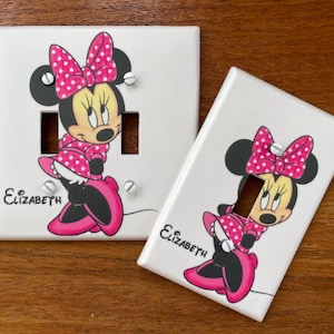 Puede incluir: Dos tapas de interruptor de luz blancas con un diseño de Minnie Mouse rosa y blanco. El nombre "Elizabeth" está escrito en cada tapa.
