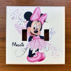 May include: A square white double light switch cover plate featuring a graphic of Minnie Mouse wearing a pink dress and bow, surrounded by purple and pink confetti. The name Maddie is printed in black script on the left side of the plate.