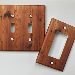May include: Two wooden-look light switch covers. One cover has two switch openings, and the other has one. The covers are brown with a wood grain pattern and are secured with silver screws. The design gives the appearance of real wood.