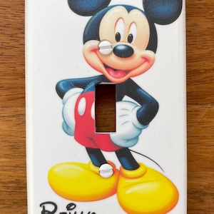 May include: White light switch cover with a colorful image of Mickey Mouse. The image shows Mickey Mouse standing with his hands on his hips, wearing red shorts and yellow shoes. The text "Reilly" is printed in black at the bottom of the cover.