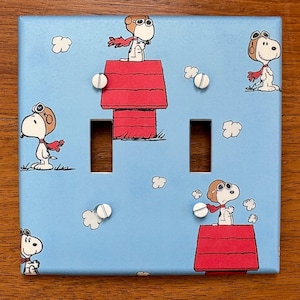 May include: A light blue double light switch cover featuring Snoopy in various poses, wearing a red scarf and aviator goggles. Snoopy is depicted on top of his red doghouse and among white cloud illustrations.
