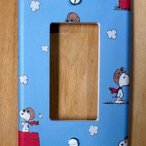 May include: A light blue wall plate featuring Snoopy in aviator gear, red scarf, and white clouds. The plate has a rectangular opening for a light switch and two white screws. The design is a cartoon theme.
