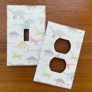 May include: Two white light switch and outlet covers with a dinosaur pattern. The pattern features watercolor dinosaurs in shades of pink, blue, green, and yellow on a white background.