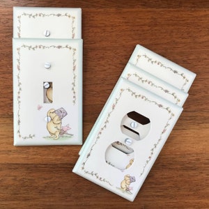 May include: Set of four Winnie the Pooh themed light switch and outlet covers. The covers are white with a green border and feature a floral design. The switch covers feature Winnie the Pooh holding a honey pot and the outlet covers feature Winnie the Pooh holding a honey pot and a bee.