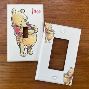 Puede incluir: Dos cubiertas blancas para interruptores de luz con una ilustración de dibujos animados de Winnie the Pooh. Una cubierta tiene el nombre "Lace" en escritura roja. La otra cubierta tiene un recorte para el interruptor y una ilustración más pequeña de Pooh.