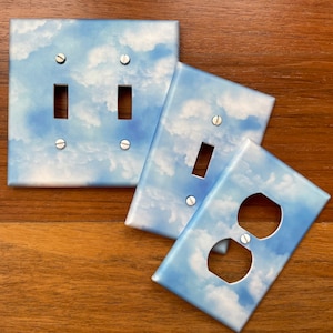 May include: Three light switch and outlet covers with a blue sky and white clouds design. The covers are made of plastic and have a glossy finish.