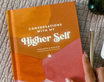 Può includere: Un diario rosa e arancione con il titolo "Conversations with my Higher Self" e le parole "Thoughts & Musings" stampate sotto. Nell'immagine si vede anche un cristallo verde e una penna argentata.