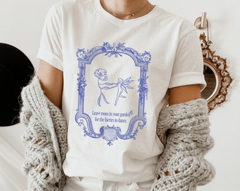May include: White t-shirt with a blue graphic of a fairy dancing in a garden. The graphic is framed in a blue ornate frame. The text "Leave room in your garden for the fairies to dance." is printed below the graphic.