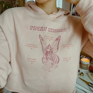 Könnte beinhalten: Hellrosa Kapuzenpullover mit dem Text "Fairy Energy" und einer Fee mit Schmetterlingsflügeln. Zusätzlicher Text beinhaltet "stop & speak with the flowers" und "feel the magic all around you."