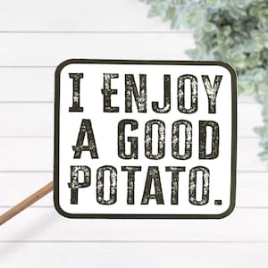 Puede incluir: Un letrero rectangular con esquinas redondeadas, con el texto "I ENJOY A GOOD POTATO." en letras negras desgastadas. El letrero es blanco con un borde negro y está montado en un palo de madera, ideal para la decoración.