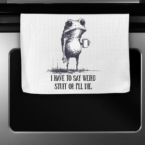 May include: White kitchen towel with a whimsical illustration of a frog holding a mug. The frog is drawn in black and white with large eyes. Text on the towel reads, "I HAVE TO SAY WEIRD STUFF OR I'LL DIE."