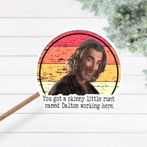 May include: A round sticker with a retro sunset design featuring a man with long hair and a beard. The text on the sticker reads "You got a skinny little runt named Dalton working here."
