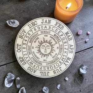 May include: A circular wooden board featuring a sun illustration at the center, surrounded by concentric rings containing the months of the year, numbers 1 through 9, letters A through Z, days of the week, and the words Yes, No, Maybe, and Rephrase.