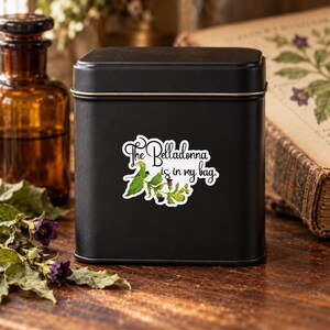 K&ouml;nnte beinhalten: Eine quadratische, mattschwarze Metalldose mit einem wei&szlig;en Aufkleber, der eine botanische Illustration aus gr&uuml;nen Bl&auml;ttern und violetten Tollkirschenbl&uuml;ten zeigt, erg&auml;nzt durch den schwarzen Schriftzug "The Belladonna is in my bag.".