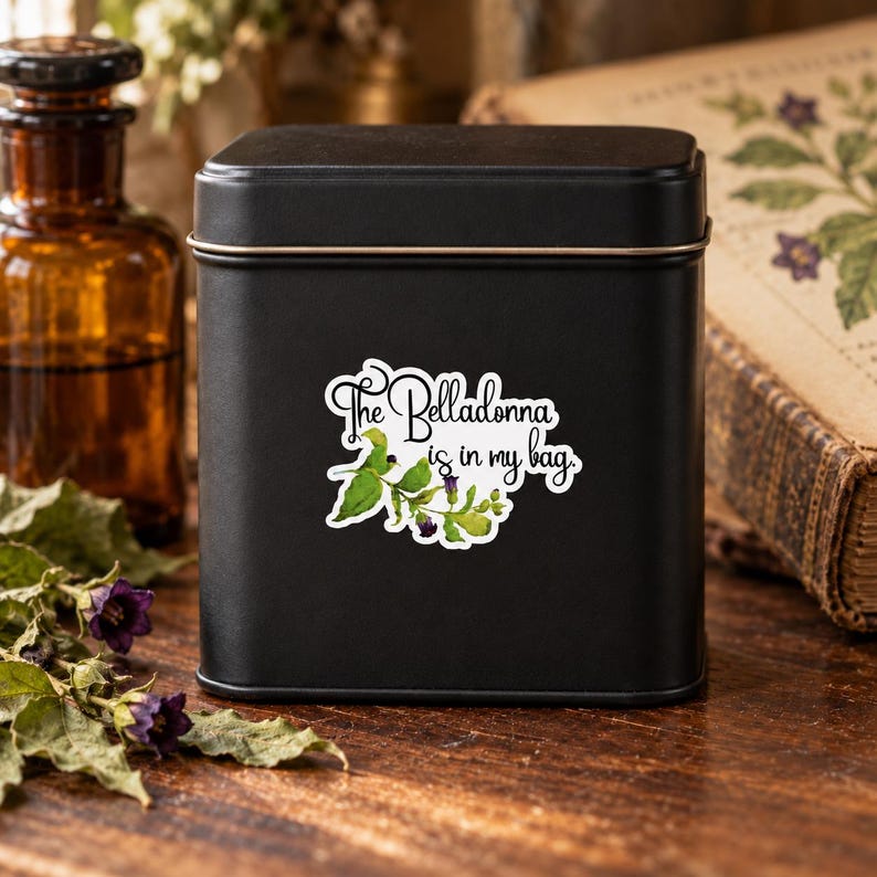 K&ouml;nnte beinhalten: Eine quadratische, mattschwarze Metalldose mit einem wei&szlig;en Aufkleber, der eine botanische Illustration aus gr&uuml;nen Bl&auml;ttern und violetten Tollkirschenbl&uuml;ten zeigt, erg&auml;nzt durch den schwarzen Schriftzug "The Belladonna is in my bag.".
