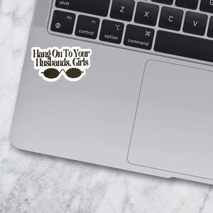 May include: A white die-cut sticker with a black border featuring the text Hang On To Your Husbands, Girls in a dark brown serif font, positioned above a graphic of dark brown oval-shaped sunglasses, applied to the corner of a silver laptop.