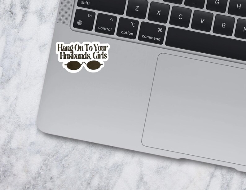 May include: A white die-cut sticker with a black border featuring the text Hang On To Your Husbands, Girls in a dark brown serif font, positioned above a graphic of dark brown oval-shaped sunglasses, applied to the corner of a silver laptop.