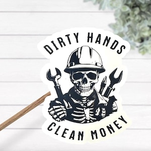 以下が含まれることがあります： ヘルメットをかぶり、レンチを持った骸骨が描かれた白黒ステッカー。「DIRTY HANDS CLEAN MONEY」の文字が骸骨の上下にプリントされています。ステッカーには白い縁取りがあります。
