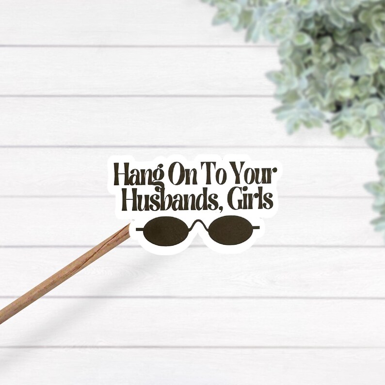 May include: A die-cut sticker featuring the dark brown text Hang On To Your Husbands, Girls above a pair of dark brown oval-shaped sunglasses. The sticker is attached to a thin wooden stick, displaying a white border around the design elements.