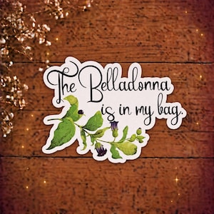 May include: A die-cut sticker featuring a botanical illustration of a belladonna plant with green leaves and dark purple flowers. The text "The Belladonna is in my bag." is written in a black, flowing script font across the white background of the sticker.