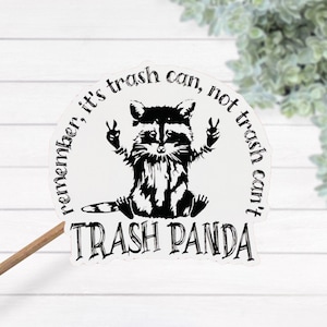 Op de afbeelding: Een witte sticker met een zwart-witte illustratie van een wasbeer met vredestekens. De tekst op de sticker luidt "Remember, it's trash can, not trash can't. Trash Panda."
