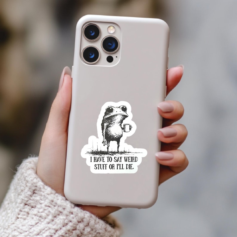 May include: A white phone case with a sticker featuring a frog holding a mug. The frog is drawn in black and white with the text "I HAVE TO SAY WEIRD STUFF OR I'LL DIE." The phone case is held by a person.