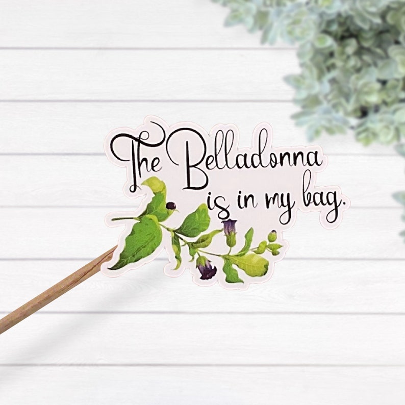 K&ouml;nnte beinhalten: Ein dekorativer Stab mit einer gestanzten Illustration einer Tollkirschenpflanze mit gr&uuml;nen Bl&auml;ttern und violetten Bl&uuml;ten. Der Text "The Belladonna is in my bag." ist in schwarzer Schreibschrift &uuml;ber dem botanischen Motiv abgedruckt.