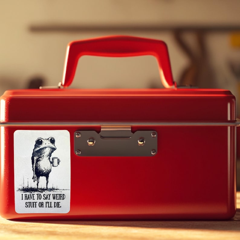 May include: A red metal lunchbox with a matching red handle. A white sticker on the front features a black and white illustration of a frog holding a mug, with the text "I HAVE TO SAY WEIRD STUFF OR I'LL DIE."