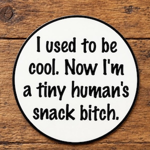 May include: A circular white coaster with a thin black border featuring the black text: I used to be cool. Now I'm a tiny human's snack bitch. The coaster has a smooth surface and is designed for placing drinks on a flat surface to protect furniture.