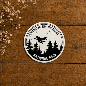 May include: A circular sticker with a white background and black ink design. The graphic features a silhouette of a figure flying on a broomstick over a forest of pine trees. The text reads FORBIDDEN FOREST NATIONAL PARK in a circular border.