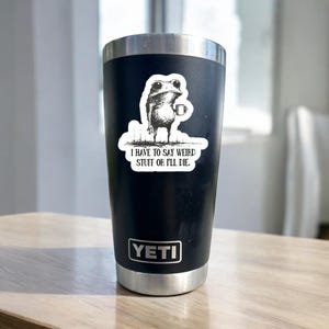 May include: Black Yeti tumbler with a white sticker featuring a frog holding a mug. The sticker has the text "I HAVE TO SAY WEIRD STUFF OR I'LL DIE." The tumbler has a silver rim and base, and the Yeti logo is visible.