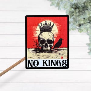 May include: A black and white skull wearing a crown with a black crow perched on a branch. The background is red with a white sun. The text "NO KINGS" is printed below the skull.
