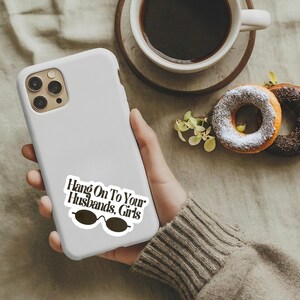 May include: A matte white smartphone case featuring a sticker with the black text 'Hang On To Your Husbands, Girls' positioned above a graphic of dark oval sunglasses. The case has a cutout for a triple-lens camera system on the upper left corner.