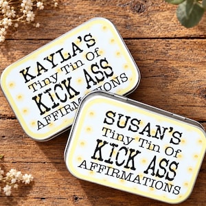 Op de afbeelding: Twee rechthoekige metalen blikjes met afgeronde hoeken, een witte achtergrond en een gele rand met stippen. De tekst op het eerste blikje luidt "KAYLA'S Tiny Tin Of KICK ASS AFFIRMATIONS" en op het tweede "SUSAN'S Tiny Tin Of KICK ASS AFFIRMATIONS" in zwarte letters.