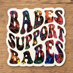 Può includere: Un adesivo quadrato con angoli arrotondati che presenta il testo BABES SUPPORT BABES in un carattere grassetto e ondulato. Le lettere sono riempite con un vivace motivo astratto multicolore di vortici rossi, blu, gialli e neri su uno sfondo color crema.