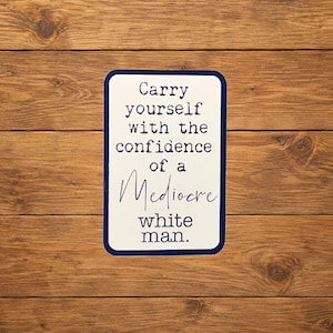 Può includere: Un cartello rettangolare bianco con un bordo blu scuro che riporta il testo nero: Carry yourself with the confidence of a Mediocre white man. Il cartello ha angoli arrotondati ed è presentato su uno sfondo di assi di legno orizzontali.