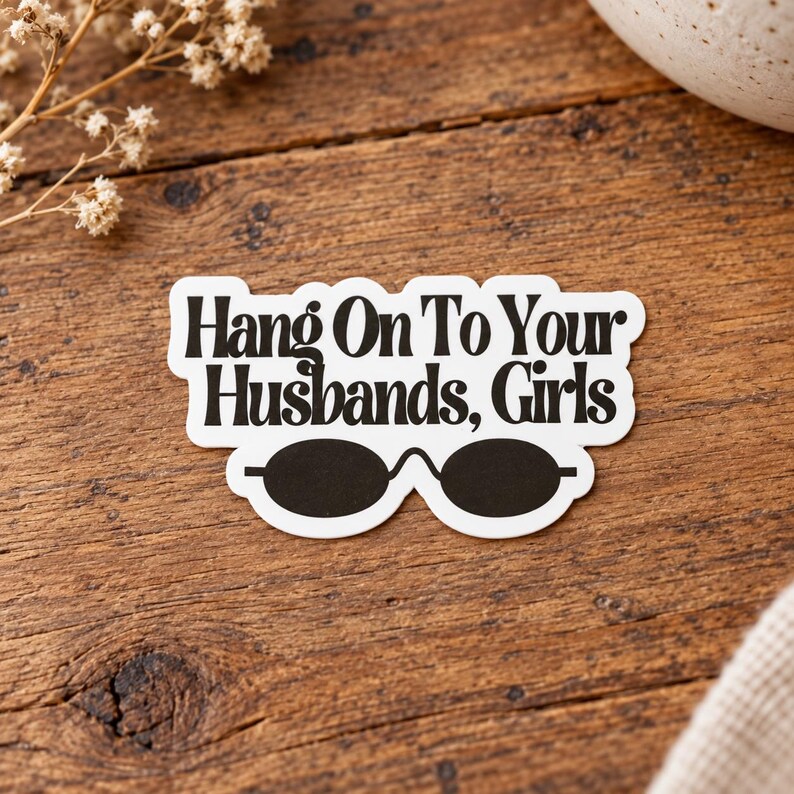 May include: A white die-cut sticker with a black border featuring the text "Hang On To Your Husbands, Girls" in a bold, serif font. Below the text is a graphic illustration of a pair of black oval-shaped sunglasses with a thin bridge.