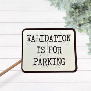 Könnte beinhalten: Ein rechteckiges Schild mit abgerundeten Ecken, weißem Hintergrund und dunkelbraunem Rand. Der Text VALIDATION IS FOR PARKING ist in einer abgenutzten, schwarzen Schreibmaschinenschrift gedruckt. Ein dünner Holzstab ist an der linken Seite befestigt.
