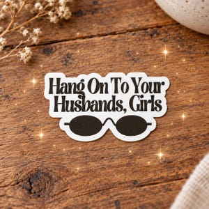 May include: A white die-cut sticker with a black border featuring the text "Hang On To Your Husbands, Girls" in a bold, dark font. Below the text is a graphic illustration of a pair of black oval-shaped sunglasses with a thin bridge.