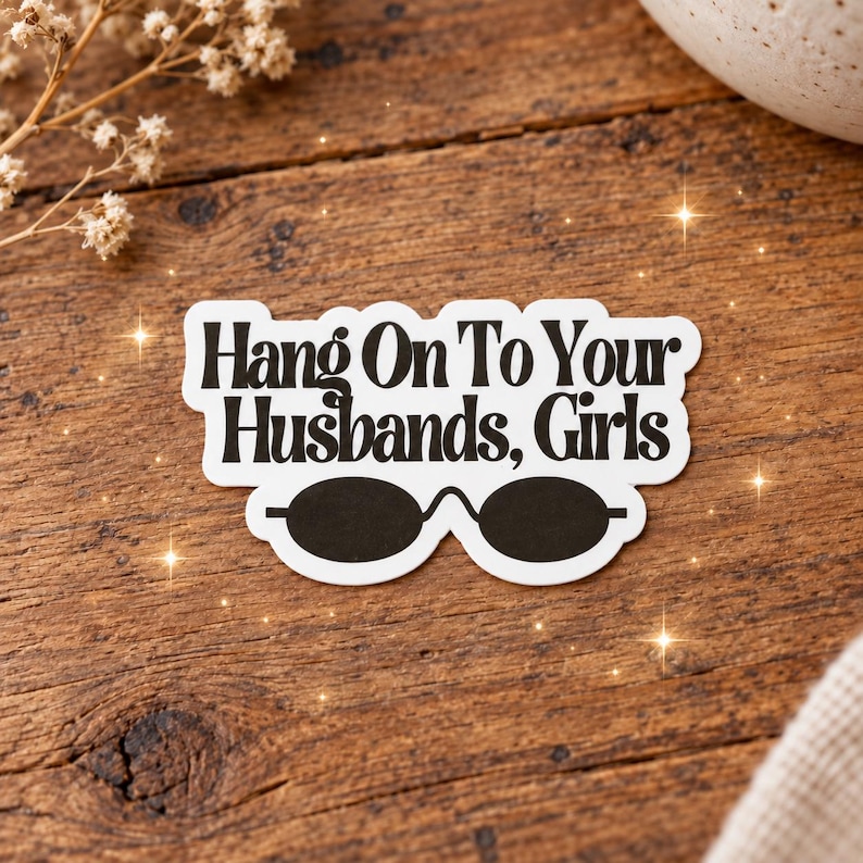 May include: A white die-cut sticker with a black border featuring the text "Hang On To Your Husbands, Girls" in a bold, dark font. Below the text is a graphic illustration of a pair of black oval-shaped sunglasses with a thin bridge.