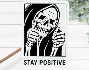 May include: Black and white sticker featuring a cartoon skeleton wearing a hooded robe and giving a thumbs up with both hands. The text "STAY POSITIVE" is printed below the image.