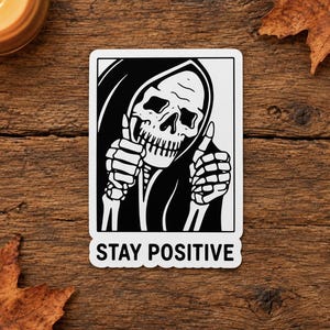 Op de afbeelding: Een rechthoekige sticker met een zwart-witte illustratie van een skelet in een mantel met capuchon. Het skelet steekt met beide handen een duim omhoog. Onder de illustratie staat de tekst STAY POSITIVE in zwarte hoofdletters gedrukt.