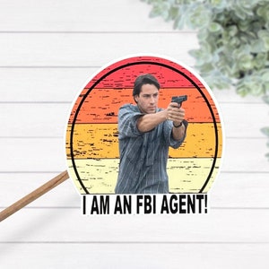 May include: A retro-style sticker featuring a man holding a gun with the text "I AM AN FBI AGENT!" in a white circle with a red, orange, and yellow sunset background.