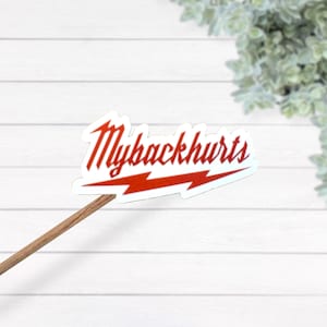 Puede incluir: Una pegatina blanca con el texto "Mybackhurts" en escritura roja, subrayada por un diseño de rayo rojo. La pegatina está adherida a un palo de madera, sobre un fondo de madera blanca.